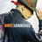 A pixelated image of a person wearing a green cap with red brim, dark jacket over white shirt, and orange accents on the cap.