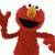 Red puppet character with large nose and wide smile, waving one arm against white background.