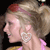 Pixelated profile of a person with light brown hair tied in a high ponytail, wearing a pink top and large white earrings against a dark background.