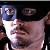 Pixelated still image shows a person with light skin wearing a white shirt and dark cap, holding a blue cylindrical object near their face against a dark background.