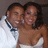 Two people in formal attire pose closely together, the person on the left wearing a white shirt with a dark blue bow tie and the person on the right in a white strapless dress, all against a dark background.