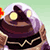 Pixelated static icon of a character with purple hair and glowing yellow eyes wearing dark clothing, set against a light green background with partial red and orange elements on the left edge.
