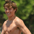 A shirtless person with light brown hair stands outdoors against a green foliage background, smiling with eyes partially closed.