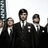 Group of men in dark suits with white shirts and ties standing against a light background, next to green with pixelated text