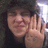A person with dark hair and a black cap holds up four fingers in front of their face, smiling slightly while wearing a dark shirt.