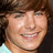 Pixelated close-up of a smiling person with brown hair and light skin, wearing a light-colored top against a dark background.