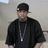 Pixelated static image of a person wearing a black cap, black jacket, and dark shirt with a lanyard and badge, standing against a light gray background.