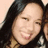 A pixelated close-up of a person with long dark hair smiling, wearing a checkered shirt against a light background.