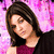A woman with shoulder-length brown hair wears a dark sleeveless top over a maroon shirt against a pink floral background.