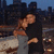 Two figures embrace on a city rooftop at dusk, with a blurred skyline and lights in the background.