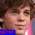 Pixelated close-up of a person with brown hair and light skin, wearing a blue rectangular graphic with red text at the bottom left.