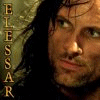 A close-up of a person with long dark hair and a beard, wearing a light-colored garment with vertical text on the left side.