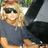 Pixelated image of a person with blonde hair wearing dark sunglasses and a dark gray short-sleeve shirt, seated with arms crossed.