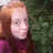 A pixelated image of a person with long red hair wearing a purple top, standing against green foliage background.