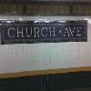 Static image of a digital street sign reading "CHURCH AVE" with pixelated text on a dark background, mounted above a blurred building facade.