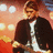 Pixelated image of a person in dark jacket and white shirt holding guitar against warm-toned background with text overlay.