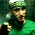 Pixelated green-toned icon of a person with dark hair and beard, wearing a cap and dark clothing against a patterned background.