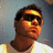 Pixelated close-up of a person with dark hair wearing dark sunglasses, light-colored shirt, and visible neck, set against a blurred background with blue and gray tones.