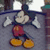 Pixelated cartoon character with large round ears, white face, red shorts, yellow shoes, and dark blue sleeves standing against a gray background.