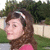 A person with brown hair and a silver headband wears a pink top, standing outdoors with green grass and a light-colored fence in the background.