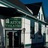 Static pixelated image of a building facade with dark roof, green and white architectural details, and dark vertical elements resembling doors or windows under a blue sky.