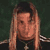 Static pixelated image of a person with long light brown hair, dark top, and visible necklace against dark green background.
