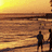 Pixelated sunset scene with two silhouetted figures standing on a beach, one figure extending an arm toward the water under warm yellow and orange sky.