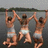 Three people in swimwear hang from a horizontal bar above water, captured across showing slight movement