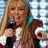 Pixelated image of a person with long blonde hair holding a microphone, set against a purple background with yellow text.