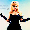 Woman with blonde hair in off-shoulder black dress and black gloves, arms extended against light blue background.