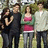 Group of five people standing outdoors in a grassy area with trees, wearing casual clothing including dark pants and various colored tops.