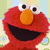 Red puppet character with large white eyes, orange nose, and wide black mouth smiling against striped background with blue and green circular patterns.