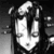 Pixelated grayscale image of a stylized figure with long hair, wearing a dark jacket over a light shirt, standing against a gradient background.