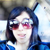 A person with shoulder-length dark hair wears large reflective sunglasses and a blue jacket with white trim, seated inside a vehicle.