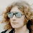 Pixelated portrait of a person with light brown curly hair wearing large reflective sunglasses and dark clothing against a pale background.