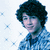 A person with dark curly hair wearing a blue patterned shirt against a light blue background with star-like patterns.