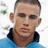 Pixelated close-up of a person with short brown hair wearing a blue shirt with a dark collar, set against a white background with faint green and gray shapes.