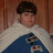 A person with dark hair wears a blue garment with white fur trim against a brown wooden background, with the word 'Jealous' in blue text at the bottom right.