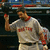 Pixelated image of a person in a white baseball uniform with red accents raising their right arm, set against dark background elements.