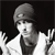 Pixelated grayscale image of a person wearing a cap and dark clothing, making a gesture with both hands raised near the face against a black background.