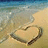 Beach scene with heart drawn in sand, waves receding, blue sky above, and a gray on the right