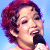 Pixelated static image of a person with curly red hair holding a black microphone against a light blue background, rendered in low-resolution 256x256 pixels.