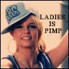 A person wearing a light blue cap with dark blue patterns, a blue and white striped sleeveless top, large hoop earrings, and a gold chain necklace, with text reading "LADIES IS PIMP" on the right side.