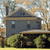 Pixelated static image of a two-story house with dark gray walls, light brown trim, and a chimney, set against a blurred background of trees and sky.