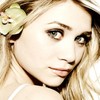A close-up portrait of a person with long blonde hair wearing a light green flower accessory on the left side of the head.