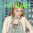 A pixelated image of a person with light brown hair wearing a white cardigan over a red top, standing against a blue background with green text above.