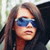 A pixelated close-up of a person with long brown hair wearing dark blue sunglasses, set against a blurred background with light and dark tones.