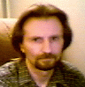 A man with brown hair and a beard sits on a brown couch, wearing a dark patterned shirt; his expression changes from neutral to smiling