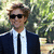 Pixelated image of a person in a dark suit, white shirt, and black tie standing outdoors with green foliage background.