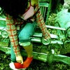 A child in a plaid shirt and teal pants sits on a green bench, adjusting red shoes with white soles.